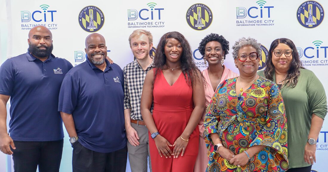 A diverse group of professionals gathers in front of a banner that reads, “Baltimore City Information Technology.”