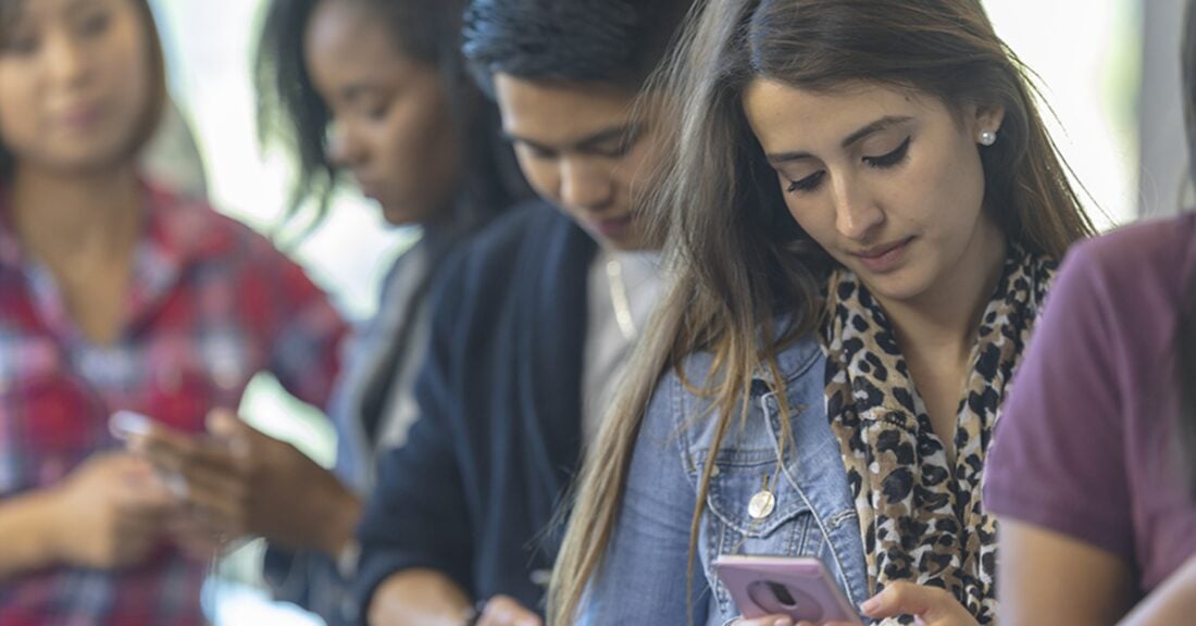 High school students of several ethnicities stand around campus and stare at their phones.