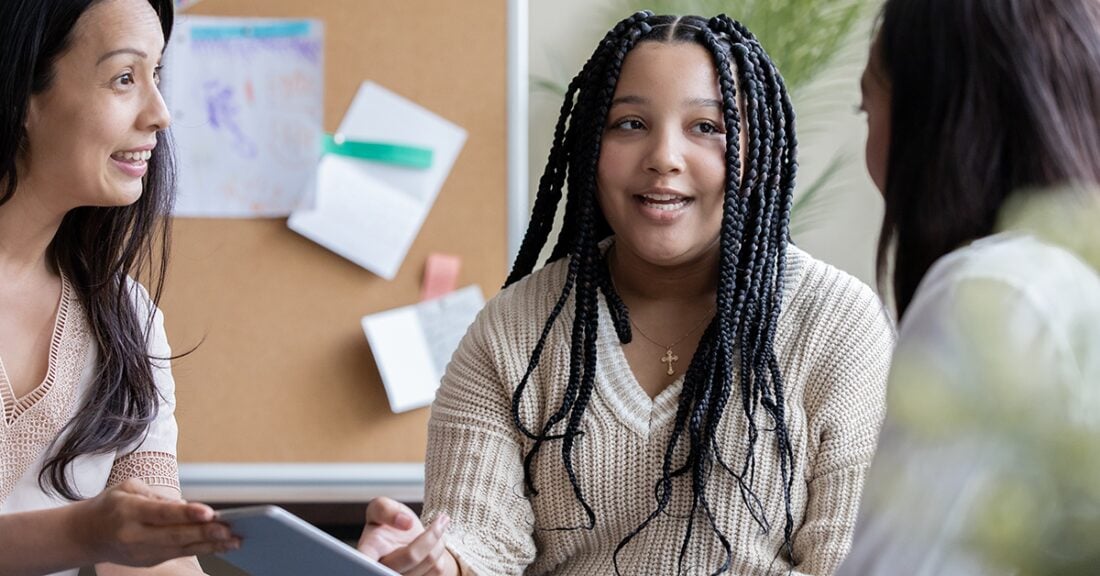 A biracial tween girl and mom meet with mature female doctor/counselor/teacher during a conference in classroom.