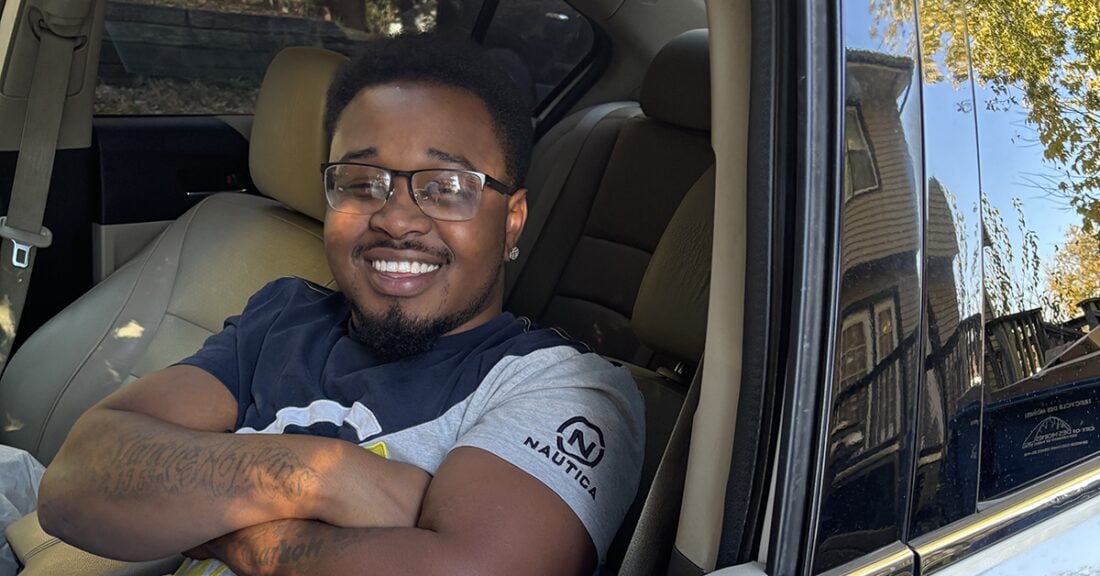 A young Black man sits in his car, smiling, with his driver's side windown rolled down ad his arms folded across his chest. the sun is shining and a house and trees are reflected in the exterior sheen of his vehicle.