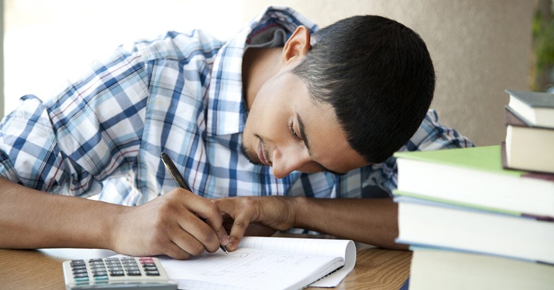 A young Latino man studies intently. His head is down with a pencil in hand and calculator at his side.