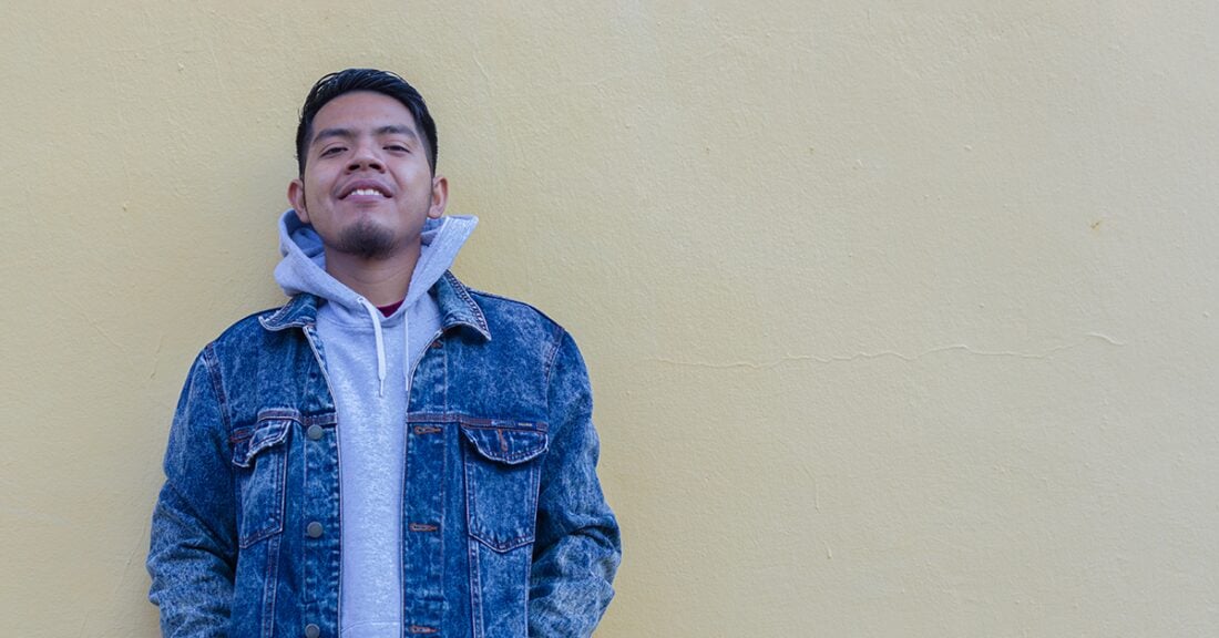 A young man in a jean jacket layered over a gray hoodie stands against a tan wall, smiling.