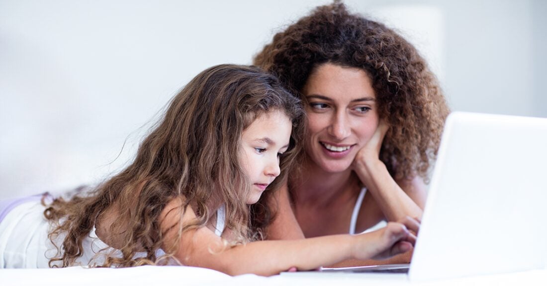 A young girl uses a laptop under the watchful eye of her mother. Both mother and daughter are laying on their stomachs; the young girl is typing on the open laptop.