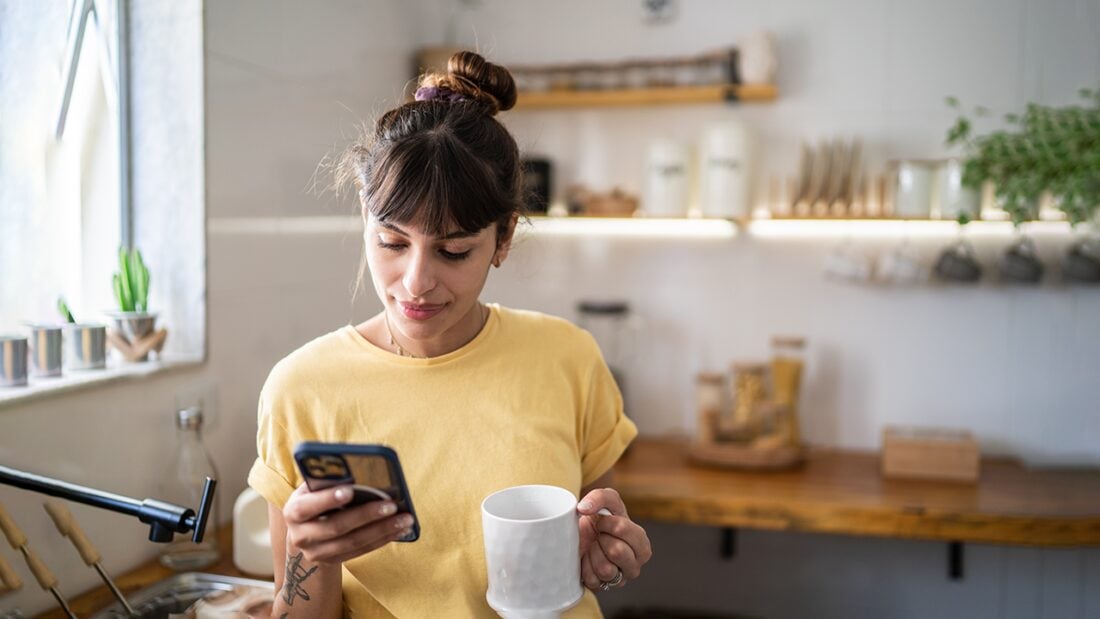 A young woman stands in a modern kitchen. She smiles as she looks at her mobile phone in one hand and holds a mug in the other.