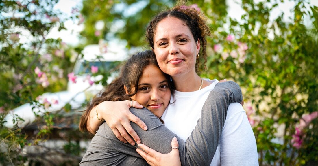 In the foreground, a Latina mother and daughter stand in a blooming garden. They embrace affectionately, smiling at the camera.