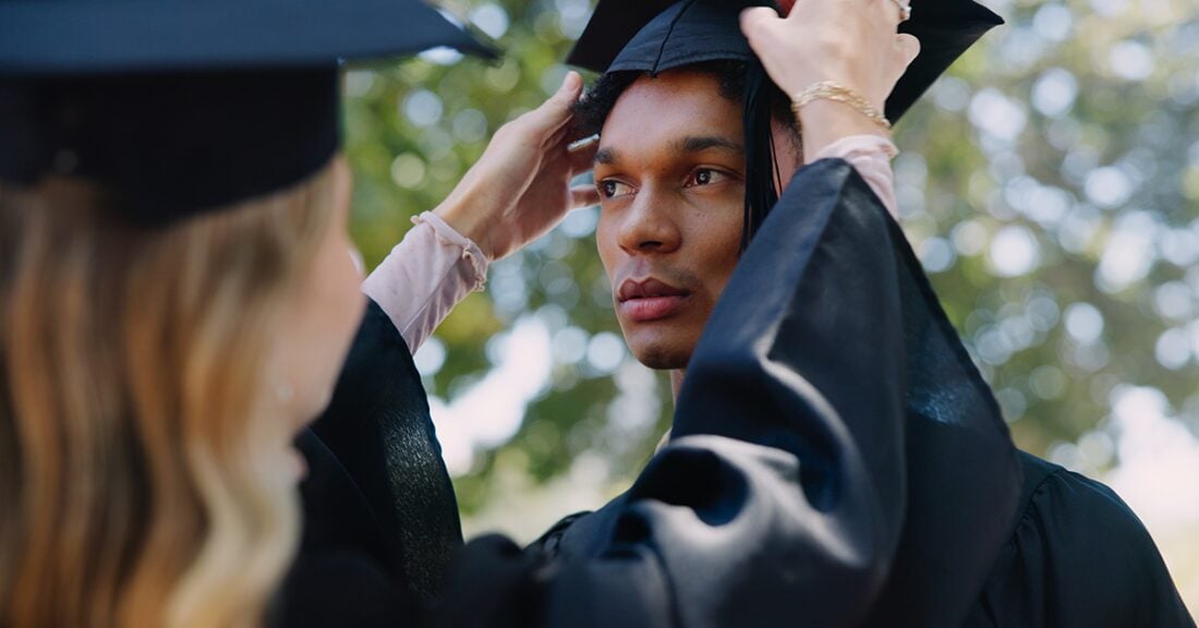 A young, Black graduate receives help adjusting his cap during a commencement ceremony.