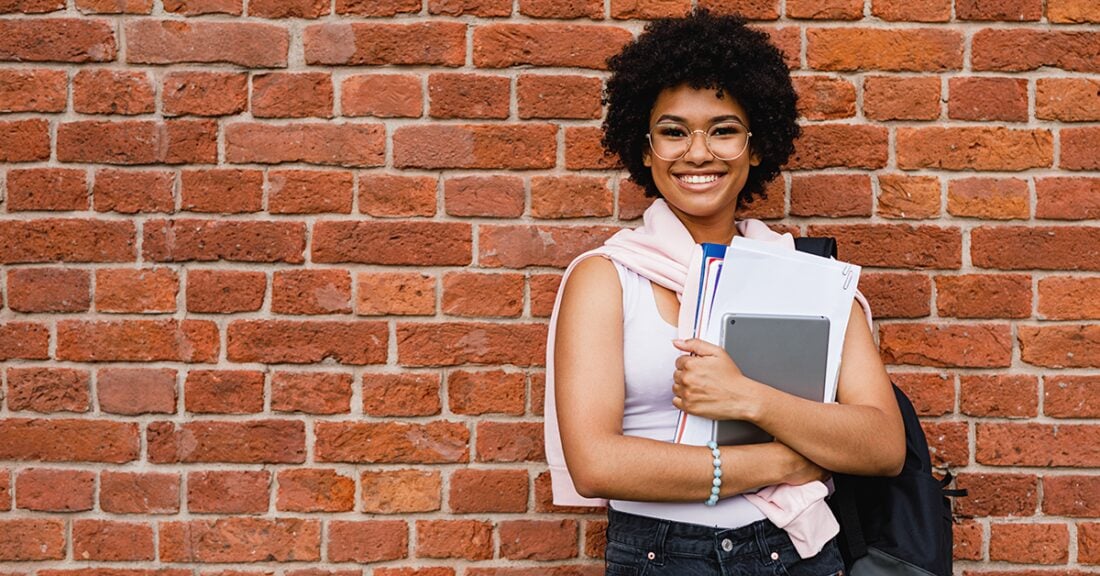 A young Black woman smiles in front of a brick wall. She holds a books in her hand and wears a backpack.