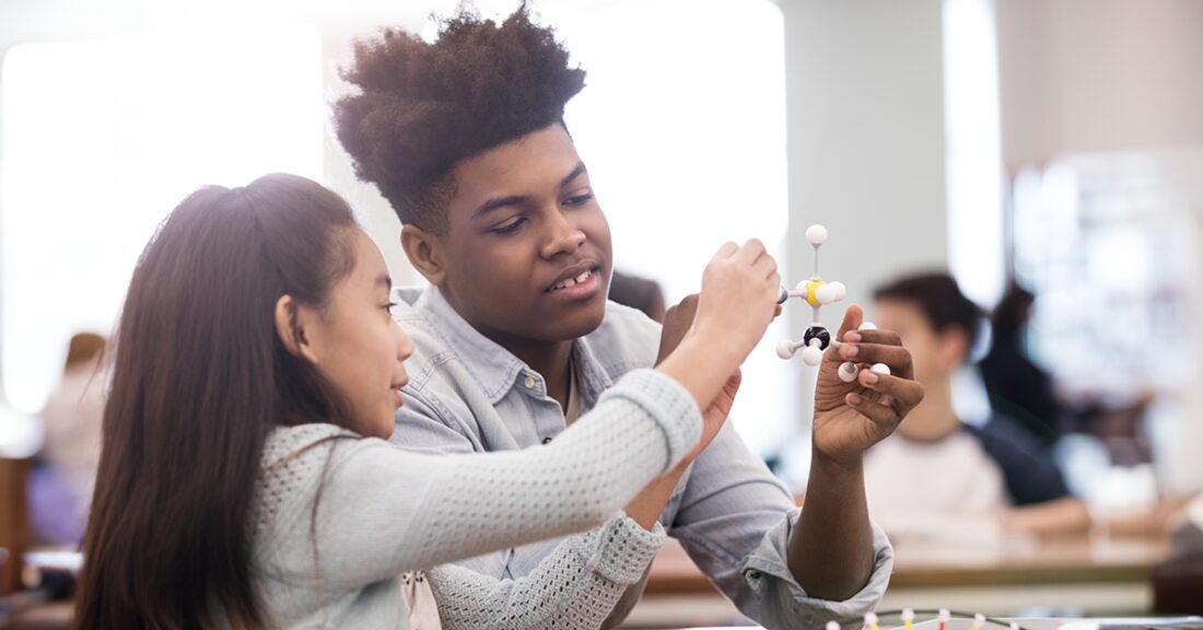 A Black teenage boy tutors a young Latin girl in science. They hold a model of a molecule and discuss it in a classroom of other students.