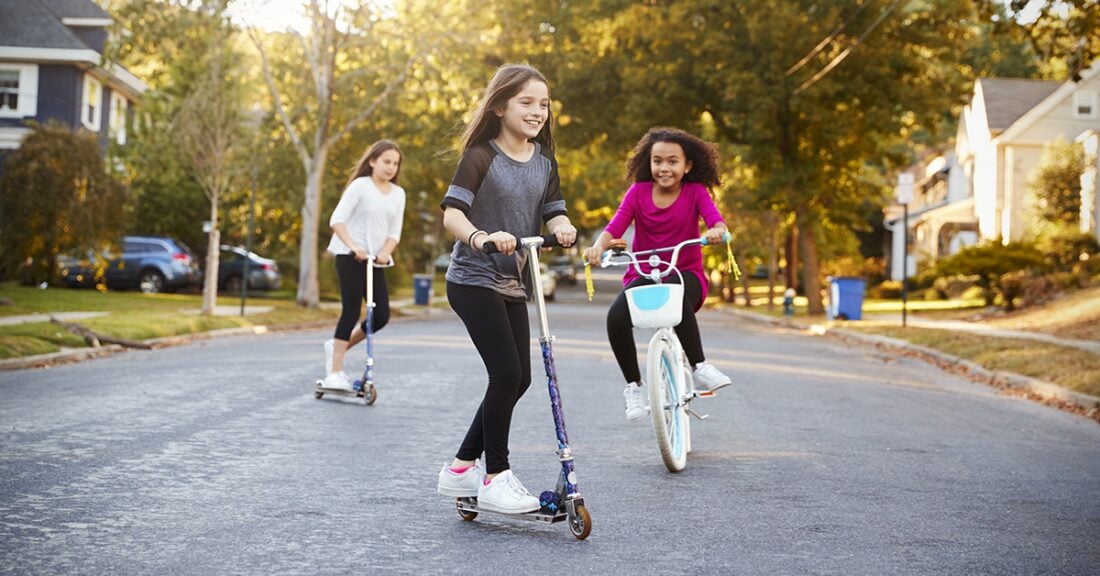 A group of three young girls scooter and bike down a sunny suburban street as the sun sets.
