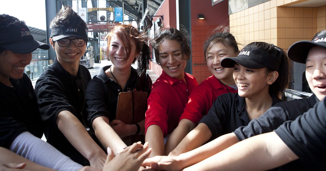 A group of young people, some wearing Juma hats or visors, stand huddled, each with an arm outstretched and their hands stacked in the center of the circle.