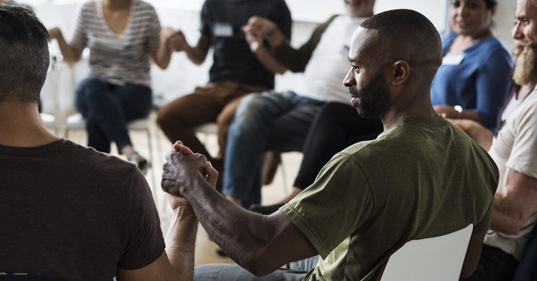 A diverse group of adults sit in a circle and hold hands in unity.