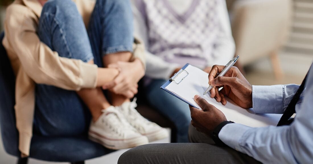A counseling session in progress. In the foreground, a professional with dark skin is holding a clipboard and pen, taking notes. Two individuals sit across from the therapist — one has knees drawn up to the chest and wears jeans and sneakers, suggesting a potentially distressed or vulnerable posture. The other wears a light sweater vest and jeans. The setting appears calm and professional.