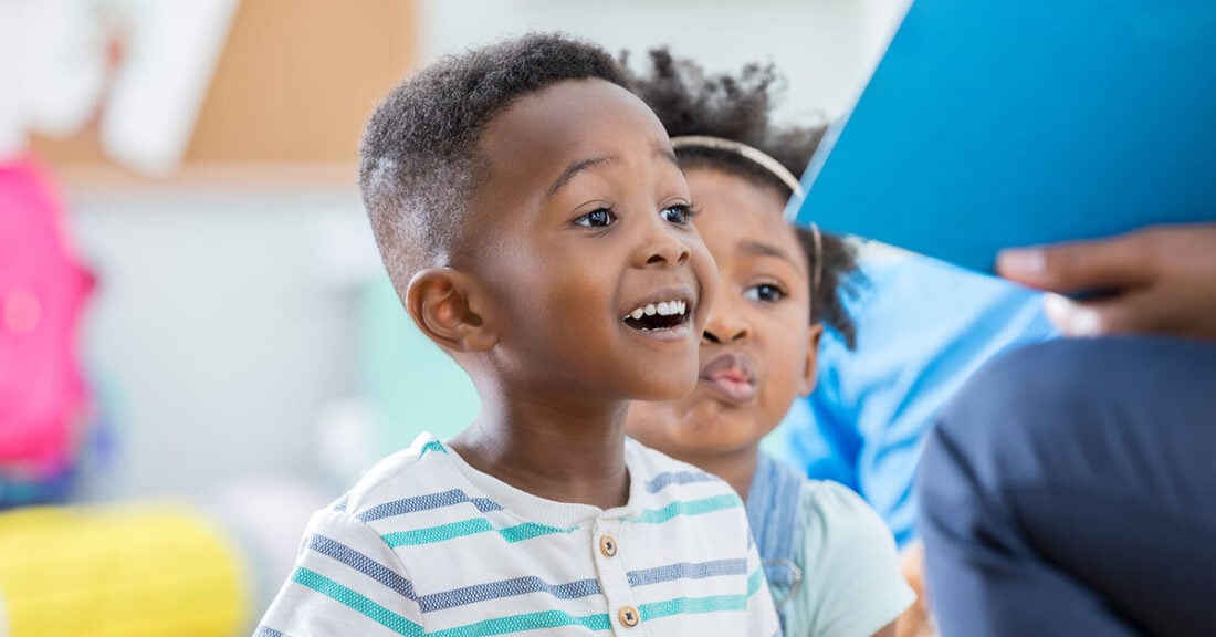 Two young children sit together, smiling and looking toward an adult holding a blue folder, as if listening to a story being read aloud.