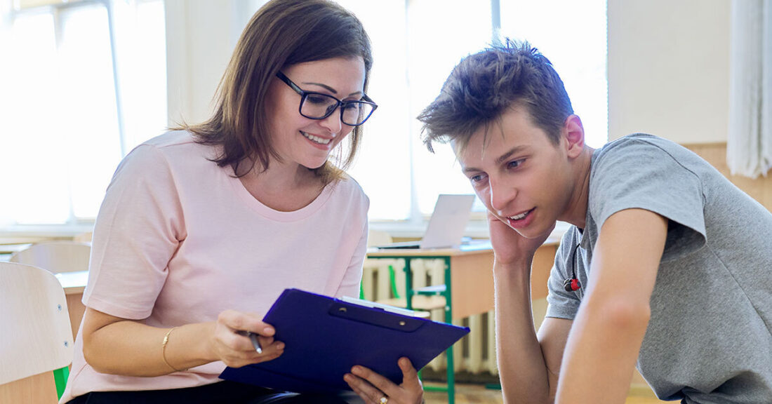 A woman with glasses and a young man smile while looking at a clipboard together in a classroom setting.