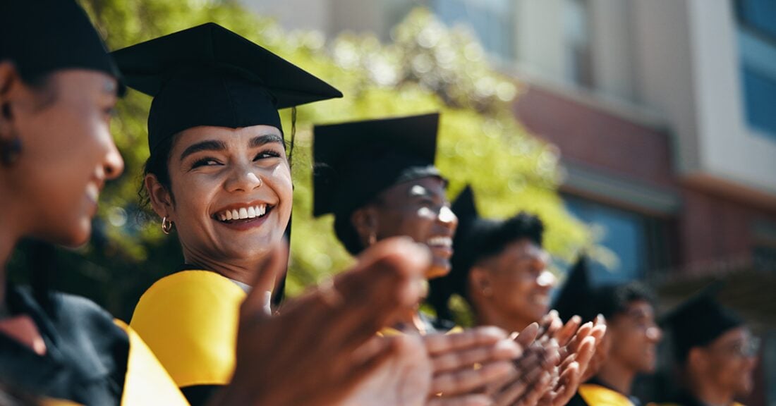 A young woman stands in a line with her high school classmates on graduation day. She is outdoors, smiling, and she and her classmates are all wearing a graduation cap and cloak.