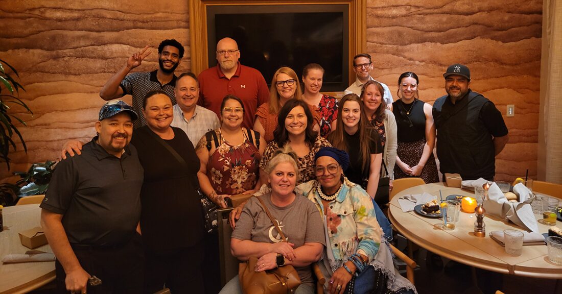 A group of sixteen people gather closely together in a warmly lit restaurant, smiling for a group photo. Some stand while others sit around a table with plates, glasses and candles, creating a relaxed and cheerful atmosphere.