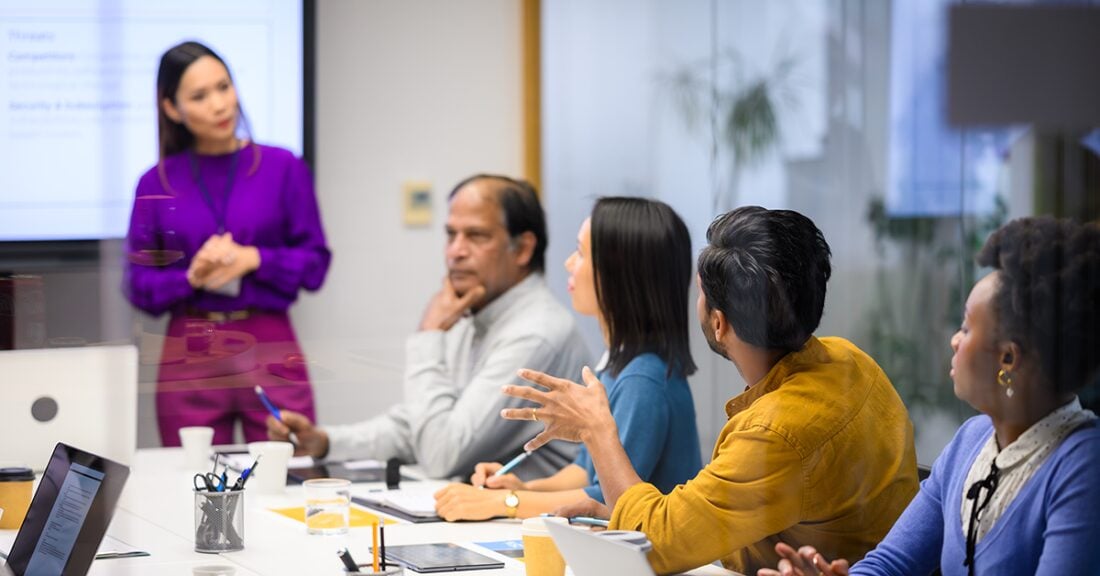 A group of professionals seated around a conference table participate in a staff meeting while a colleague presents at the front of the room.