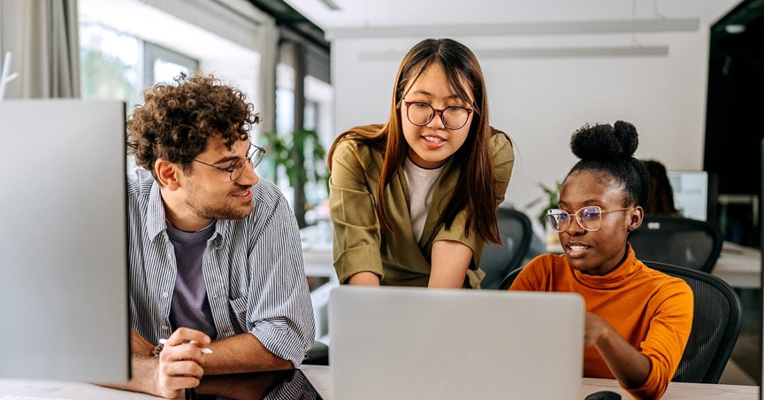Three coworkers collaborate at a desk in a bright office, looking at a laptop together — one person gestures at the screen while the others listen and engage.