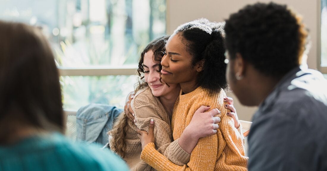 Two young women embrace during a group gathering, illustrating connection and peer support among young people.