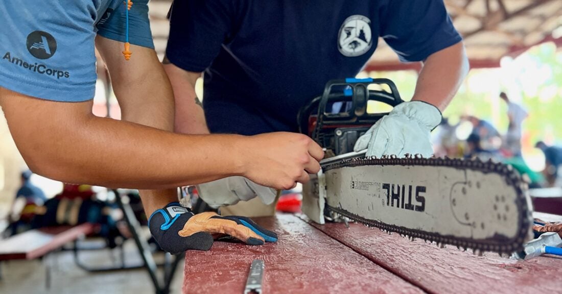 Two people work together to maintain a chainsaw on a workbench, one wearing an AmeriCorps shirt.