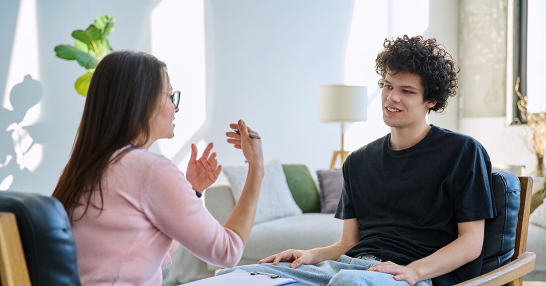A young man sits in a chair, smiling as he talks with a woman who gestures with her hands.