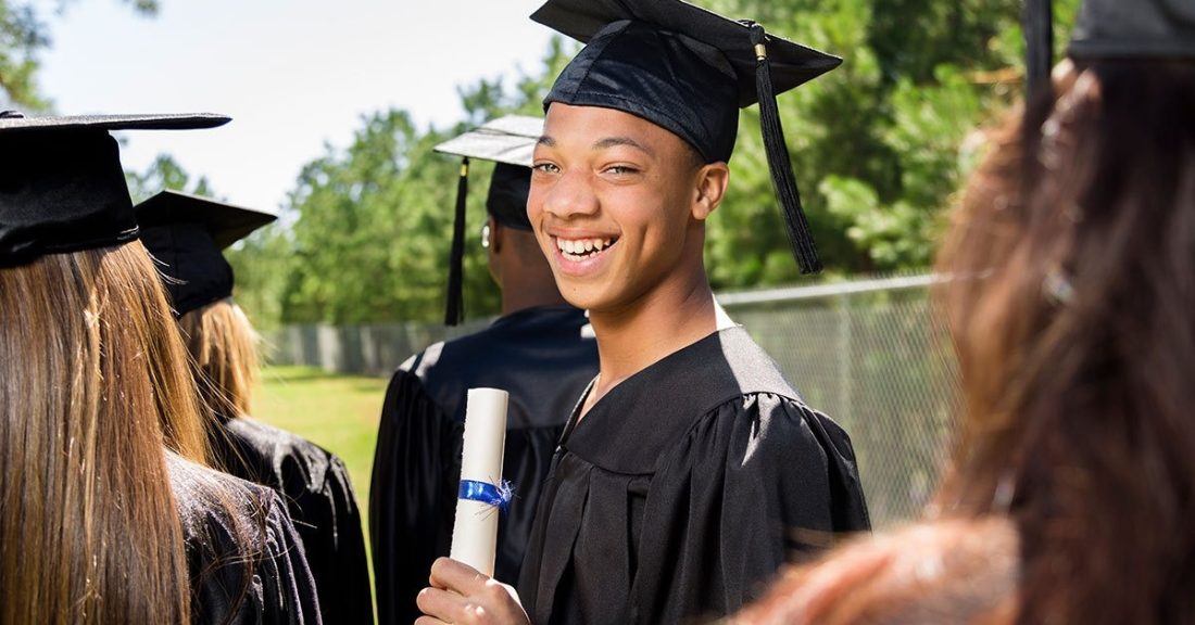 Young black man in cap and gown smiles at the camera while holding his diploma.