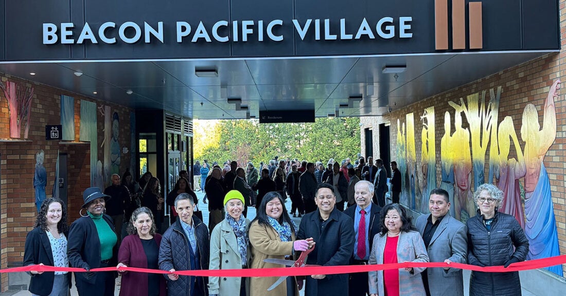 11 people stand outdoors in front of a red ribbon for ribbon cutting ceremony. The words "Beacon Pacific Village" are on a building behind them.