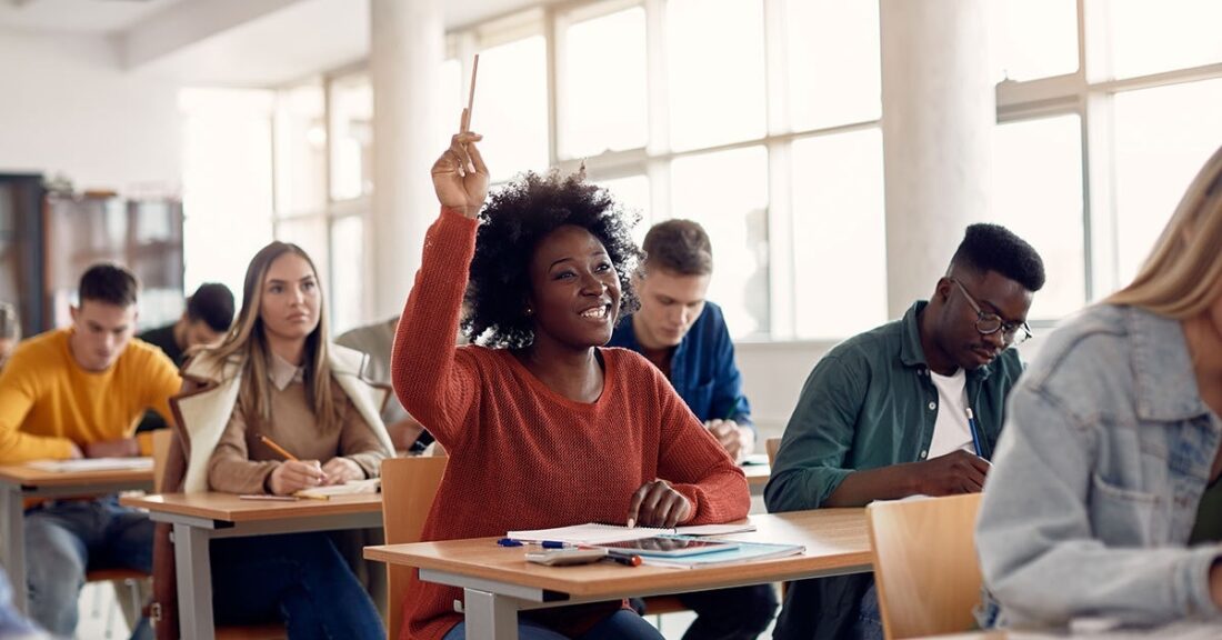 A young black woman sits among classmates, at a desk, with her hand raised.