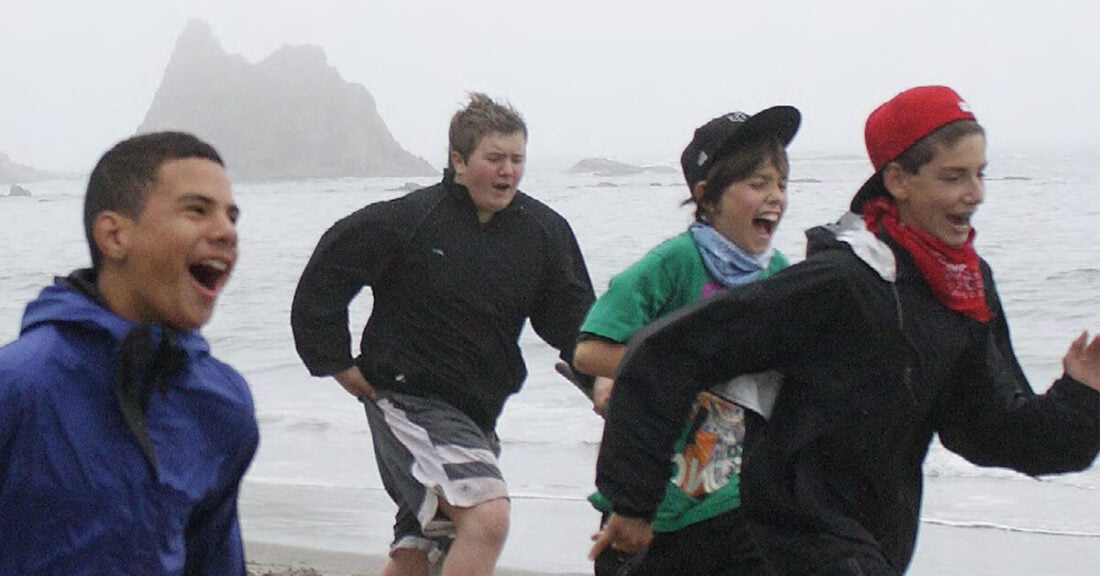 Four kids run and laugh together along a beach on a cloudy day, waves and a rocky formation visible in the background.
