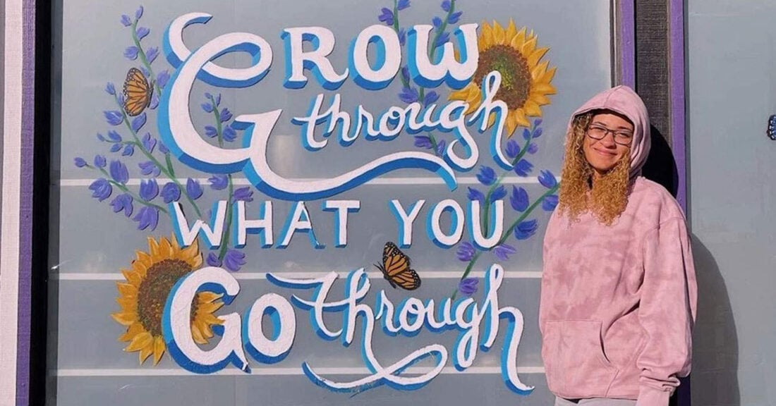 A young woman in a pink hoodie stands in front of a painted walll that says: "Grow through what you go through"