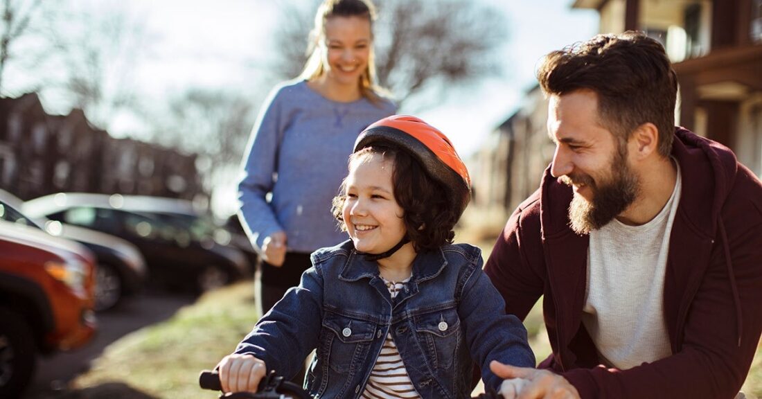 Family is outside teaching their smiing young daughter how to ride a bike