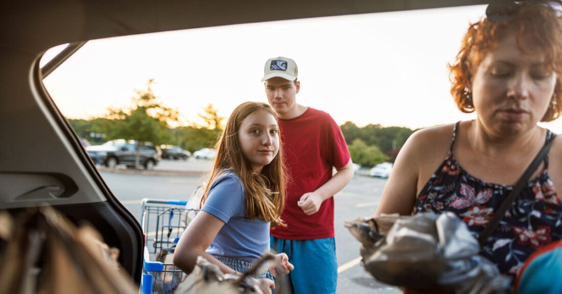 A woman and two children load groceries into the trunk of a car in a parking lot at sunset, reflecting family life and the rising cost of essentials.
