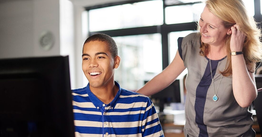 Teacher stands behind a student working at a computer screen