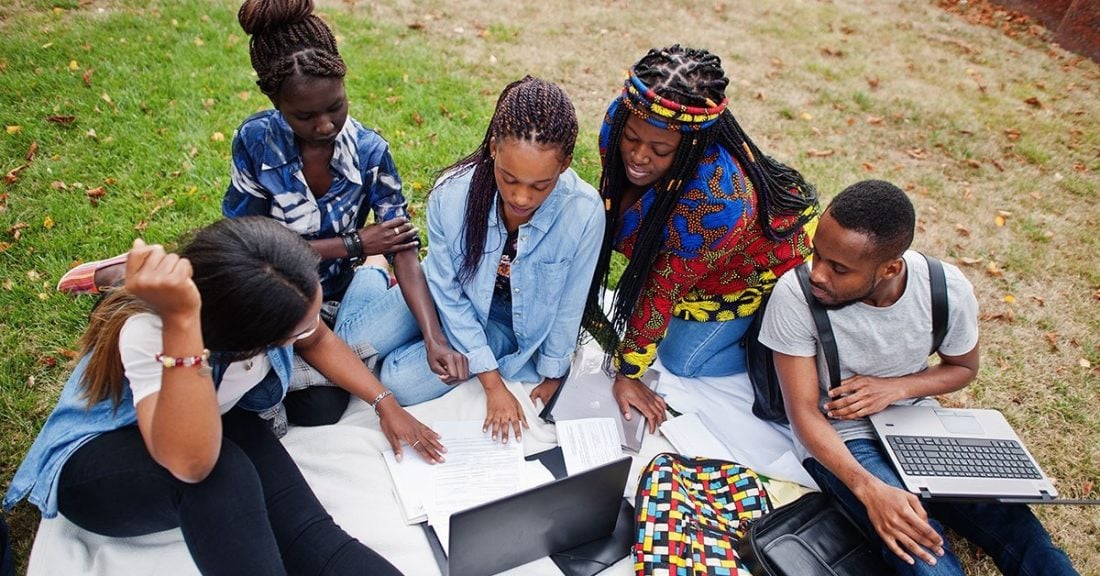 Five community college students gather on a lawn, with laptops and papers in front of them. They are seated and are reviewing both the laptop screens and papers. Four students are female, and one is male.