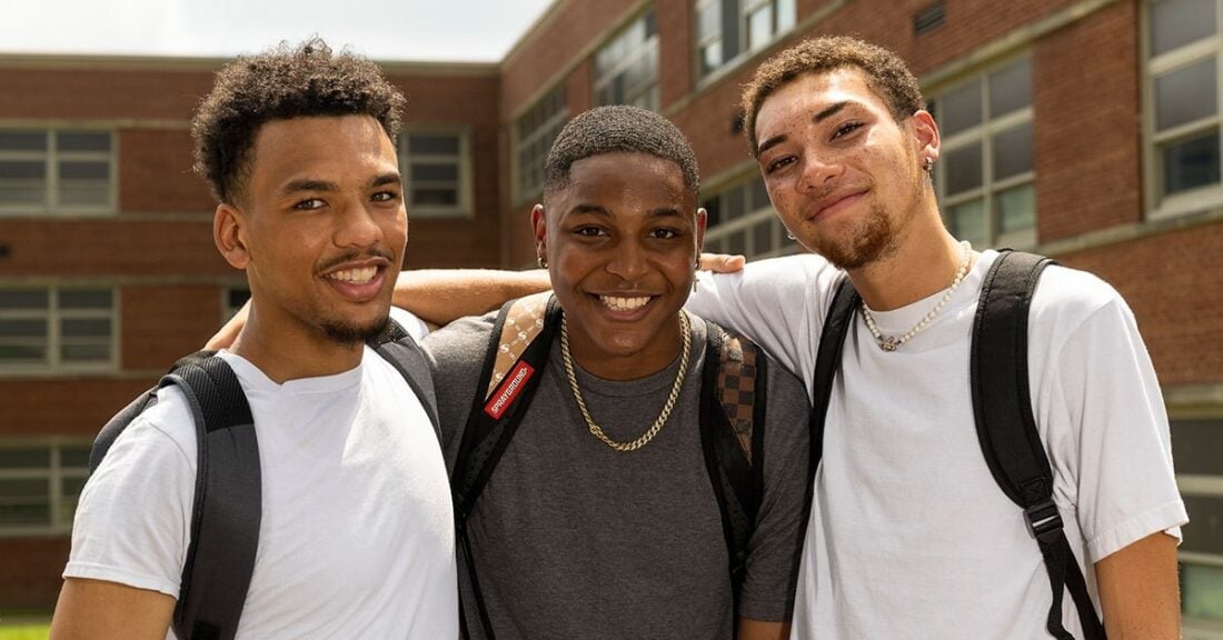 Three young black men wearing backpacks stand close together, smiling at the camera.