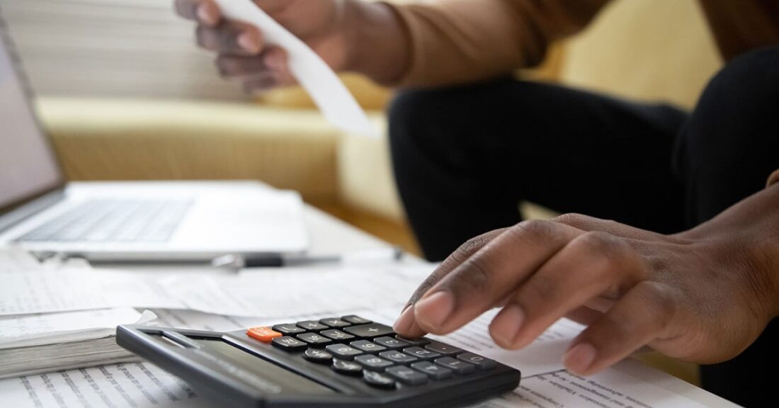 Close up of a black person sitting at a desk, in front of a laptop and papers. One hand is using a calculator