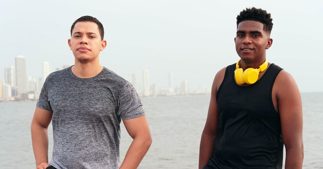 Two young adults of color. One in a gray tee shirt and another in a black tanktop and wearing yellow headphones around his neck.