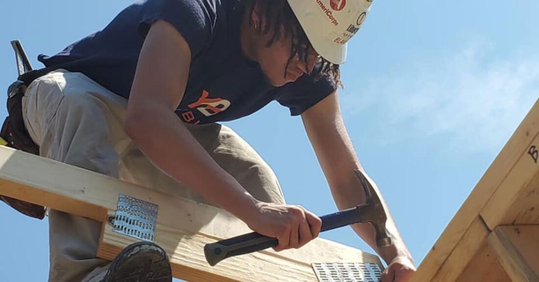 Young Black man wears a hard hat and hold a hammer; standing on the frame of an unfinished building