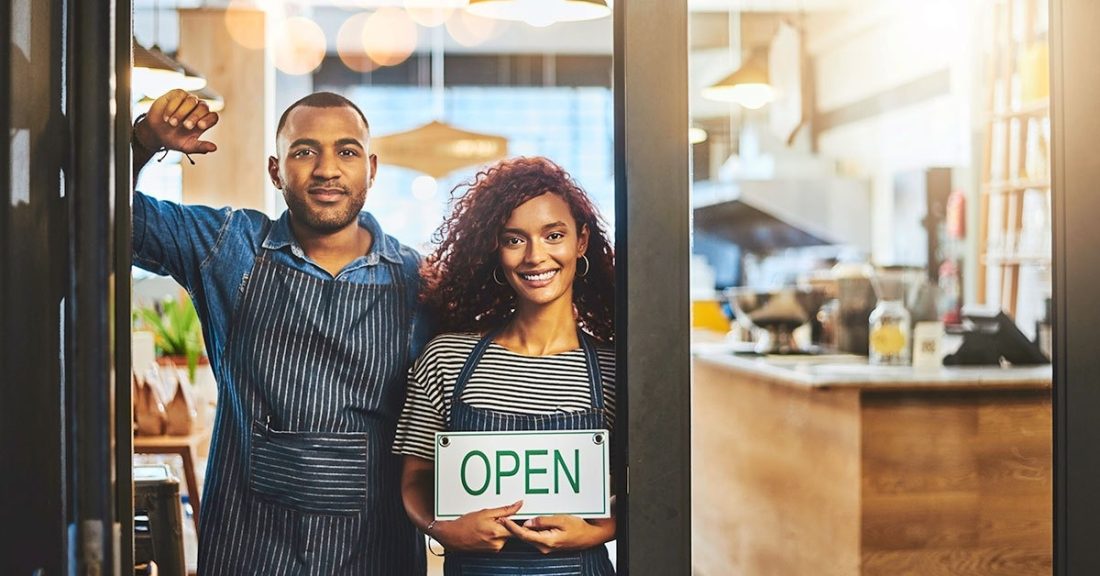 Man andA black man and a black woman stand in the doorway of a store. The woman is holding a white sign that says open. Both are smiling an wearing striped aprons.