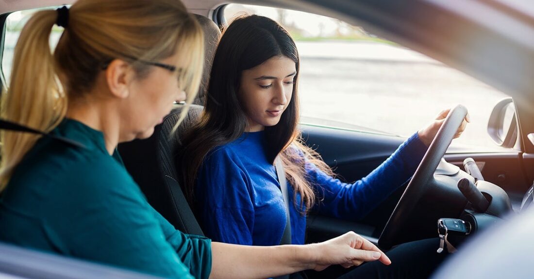 A young girl in a blue shirt and long dark hair sits behind the wheel of a vehicle. An older woman sits next to her and appears to be helping the girl learn how to drive.