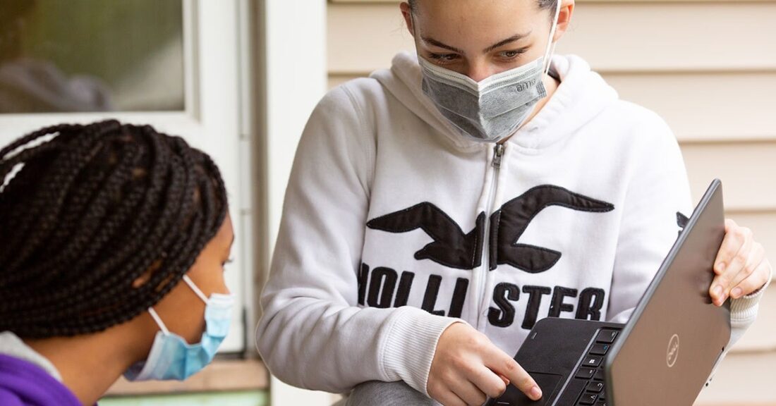 Woman shows her laptop screen to another woman. Both women are masked to prevent the spread of infection, presumably from the coronavirus.