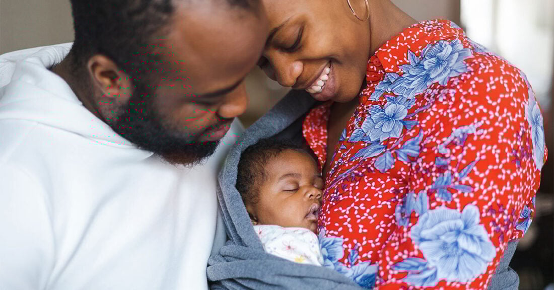A smiling Black couple lovingly holds their sleeping baby wrapped in a blanket.