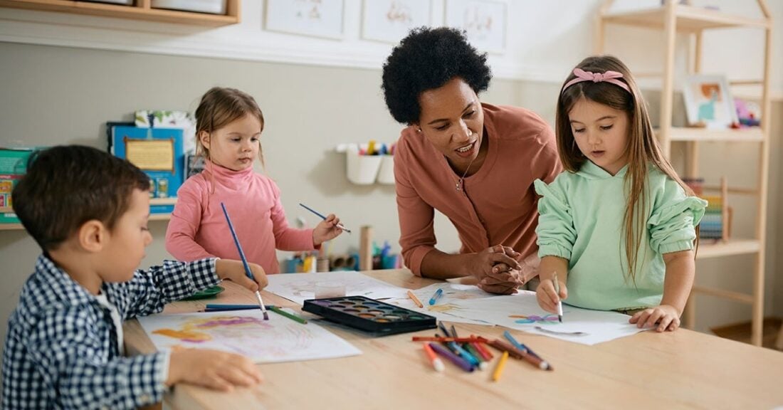 A Black female child care provider helps three young children with a craft activity