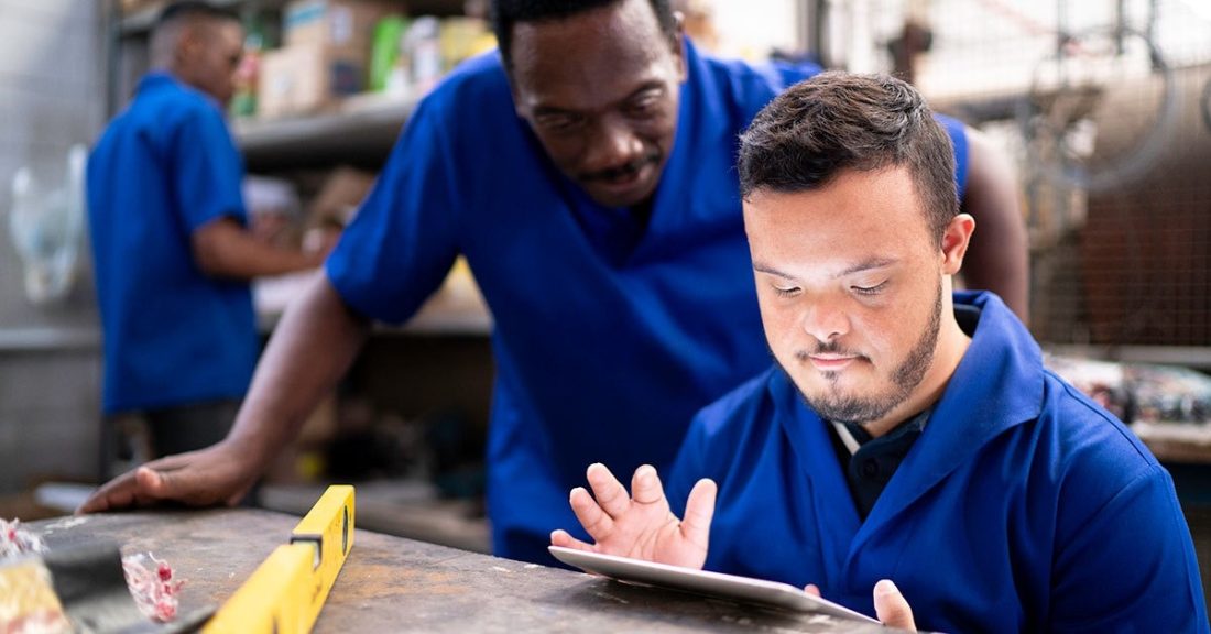 Young man with Down Syndrome works on a tablet; a man stands next to him, observing.