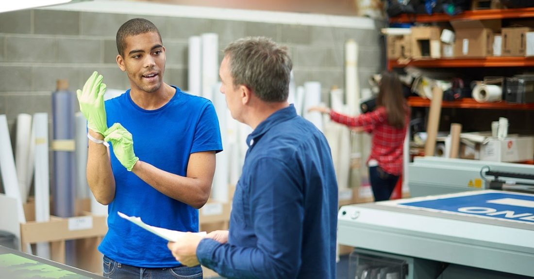 Young black man stands in a print shop, with gloves on his hands listening to an older white man give him instructions
