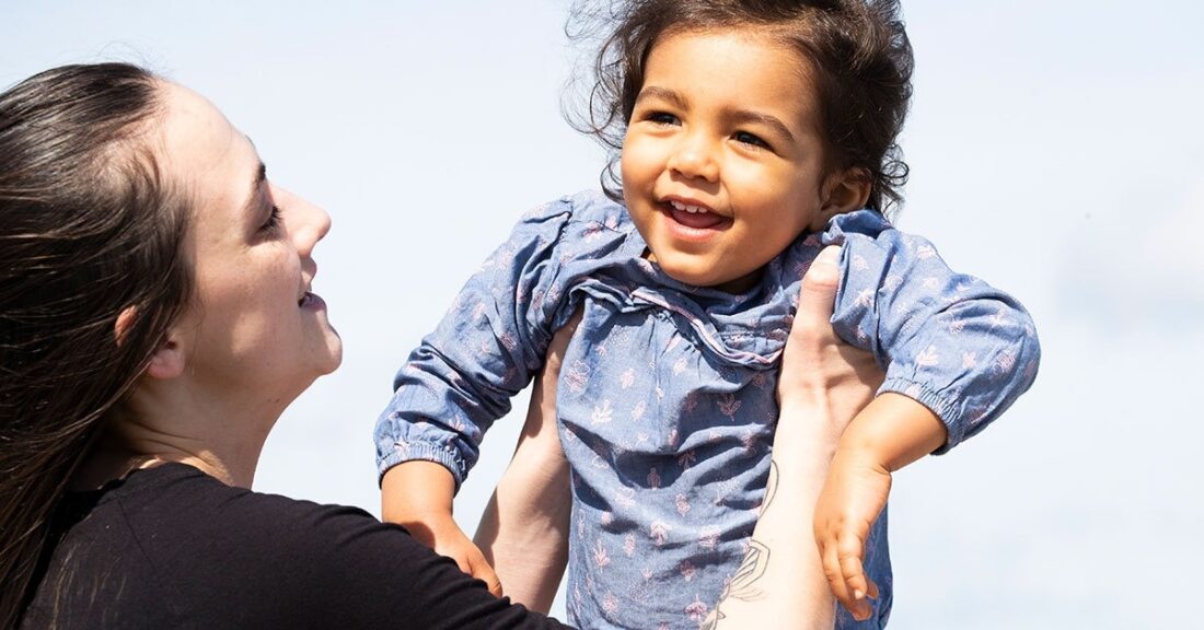 Woman with toddler. They are facing one another, the woman is holding up the child in front of her; both are smiling.