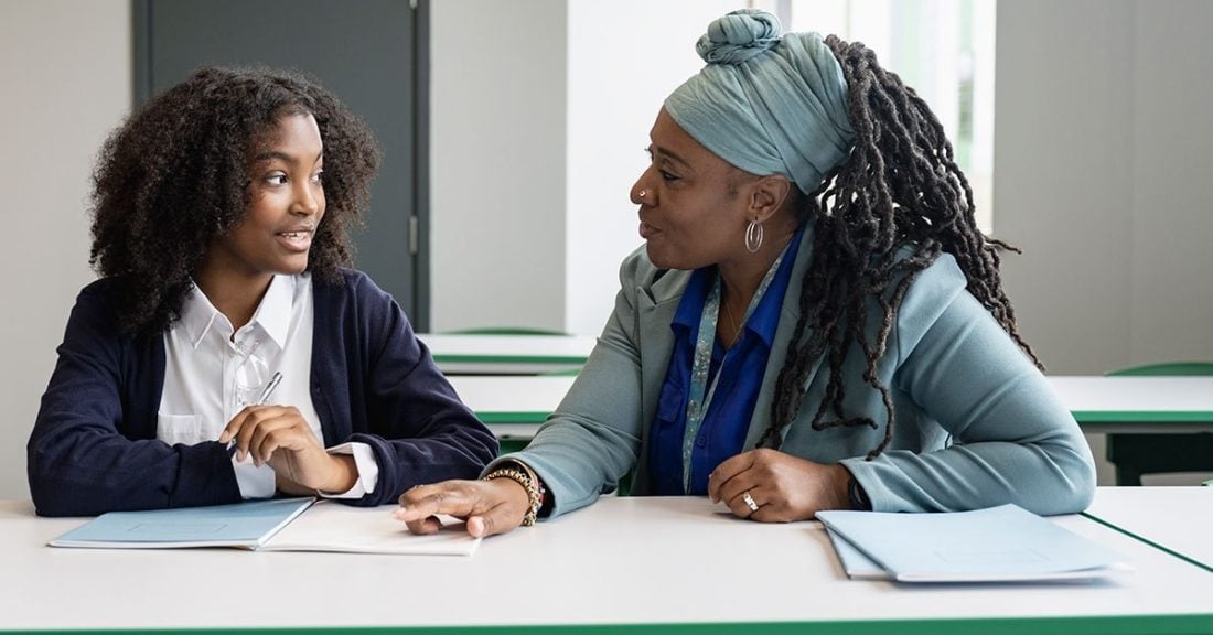 Two black women — one a young student — are sitting down next to each other and talking at a table.