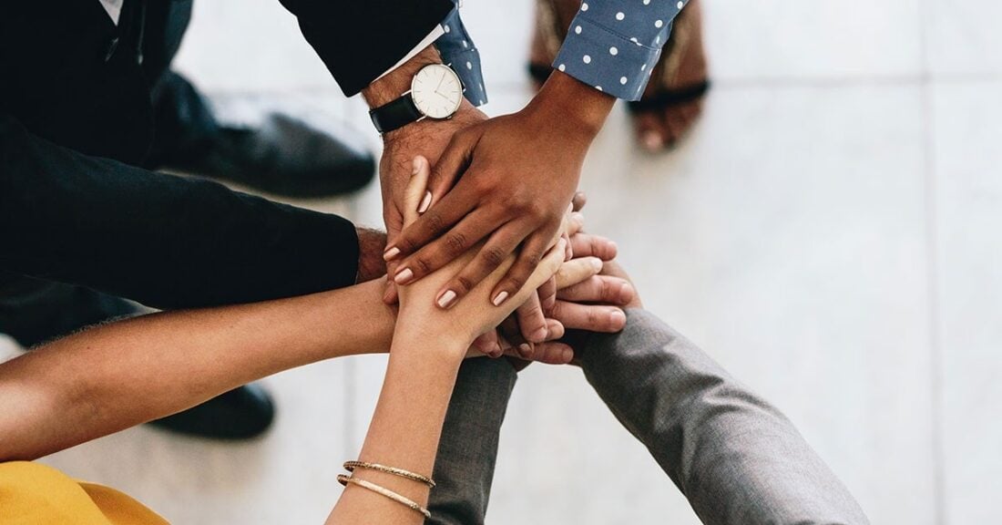 A multiracial group of young people form a circle, each putting a hand in the middle--one on top of the other.