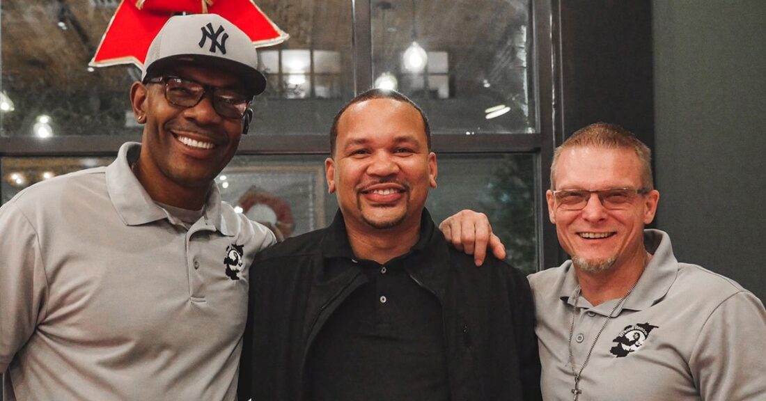 Three men stand shoulder to shoulder, looking at the camera and smiling.