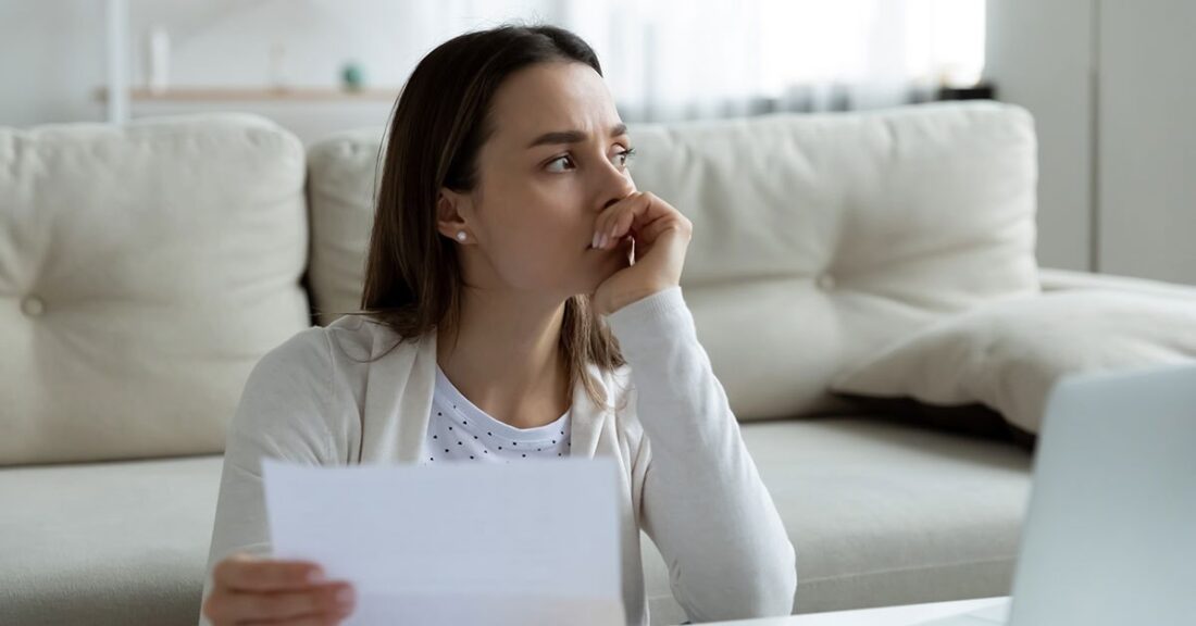 A woman holding a sheet of paper, sitting at a desk looking concerned, with a white couch behind her.